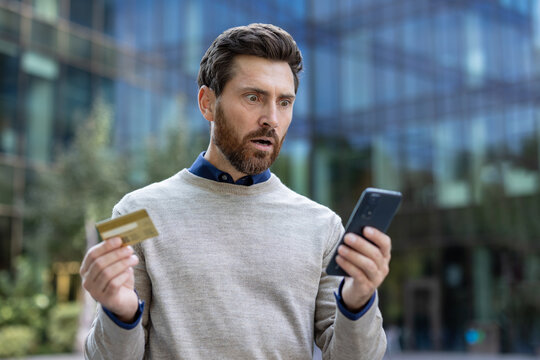 Shocked young man standing on the street, holding a bank credit card and looking at his phone in surprise