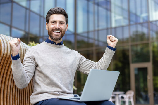 Portrait of a young happy man sitting on a bench on the street, holding a laptop on his lap, rejoicing in success