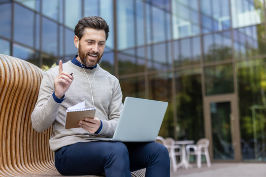 A young man is holding an online meeting on a laptop, sitting on a bench outside, talking and taking notes in a notebook