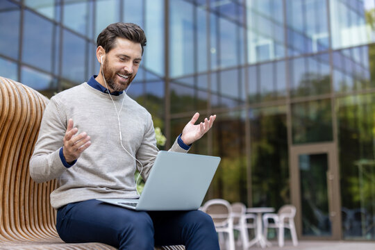 Smiling young man sitting on a bench on a fishing pole near the office and chatting on a video call on a laptop, gesturing with his hands