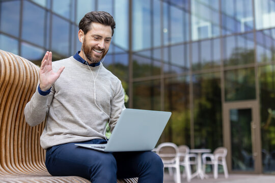 A young man is holding an online meeting on a laptop, sitting on a bench on the street, greeting the camera