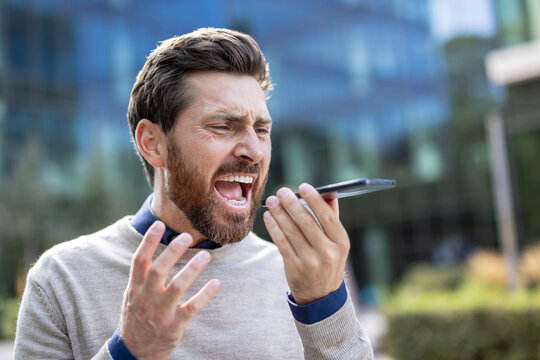 Close-up photo of an angry young man standing outside, talking and shouting into the speakerphone of the phone he is holding in his hand
