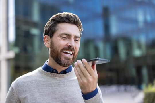 A close-up photo of a young man standing outside and talking on the speakerphone he is holding in his hand