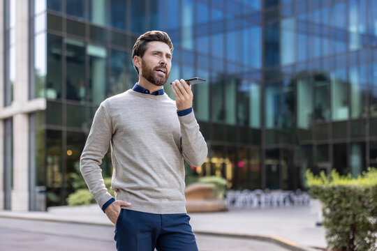 Young businessman standing on the street, holding his hand in his pocket and talking on the phone on the speakerphone