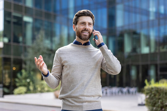 Smiling and young male businessman standing on the street near a building, talking on the phone, gesturing with his hands