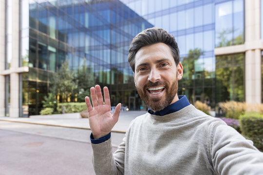 A close-up photo of a young man standing near an office building, greeting and talking to a camera he is holding in his hand