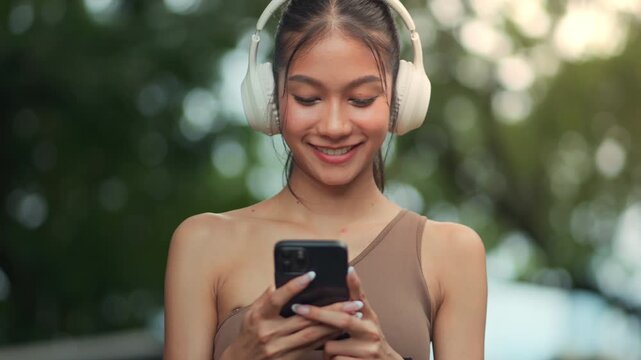 Happy young Asian woman in sportswear using smartphone and wireless headphones at park, fitness technology and active lifestyle