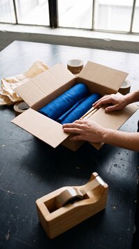 A person carefully packing blue fabric rolls into a cardboard box at a workshop desk for shipping and order fulfillment