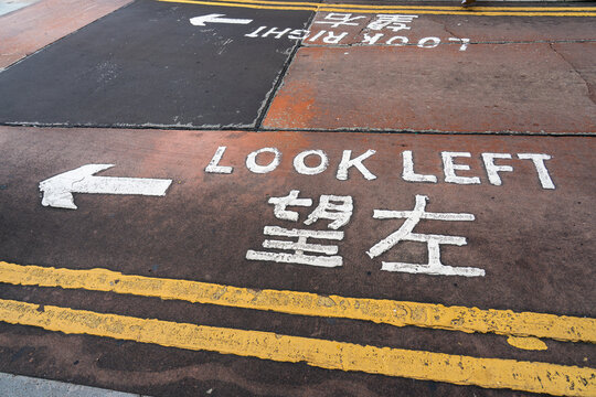 Top-down perspective of bilingual road marking Look Left and 望左 painted in bold white letters on textured asphalt with yellow lines.