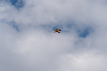 A yellow vintage biplane cuts through soft white and gray cloud formations across a bright, dramatic sky. © Sergej