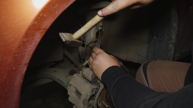 Male mechanic's hands using a wrench and hammer to loosen a bolt on the shock absorber while repairing the vehicle's suspension system in an auto repair shop or garage