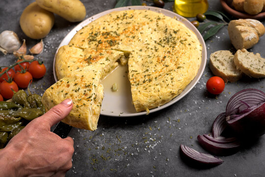 Hand taking a portion of juicy traditional Spanish omelette on a plate with bread, oil, tomatoes and peppers on dark background