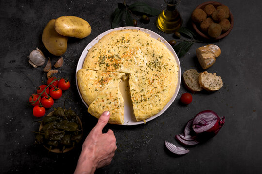 Hand taking a portion of juicy traditional Spanish omelette on a plate with bread, oil, tomatoes and peppers on dark background