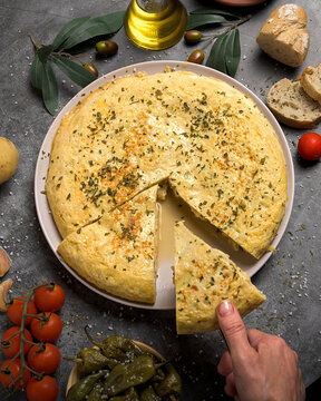 Hand taking a portion of juicy traditional Spanish omelette on a plate with bread, oil, tomatoes and peppers on dark background