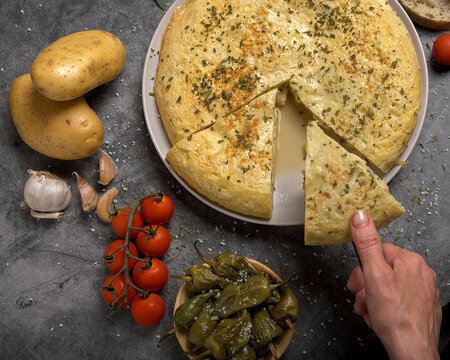 Hand taking a portion of juicy traditional Spanish omelette on a plate with bread, oil, tomatoes and peppers on dark background