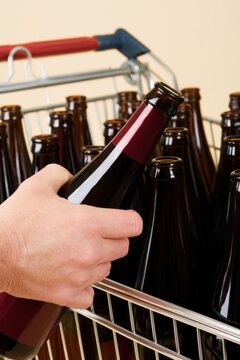 Shopper selecting dark glass beverage bottle from a metal shopping cart filled with similar bottles, hand gripping the neck with five fingers visible