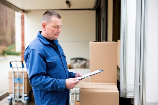 Middle-aged delivery worker in blue uniform checks shipment list beside truck in small warehouse loading area with cardboard parcels and hand trolley nearby