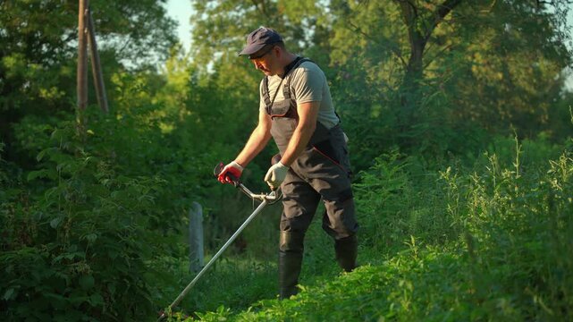 Male gardener in gray shirt and black overalls operates a string trimmer, cutting grass and weeds in a lush green garden path surrounded by trees and dense vegetation during daylight