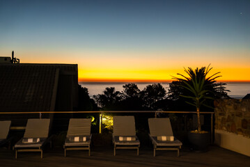 Fototapeta premium Sun loungers with folded towels line a hotel terrace facing the sea horizon during a vibrant evening sunset in Cape Town, South Africa.