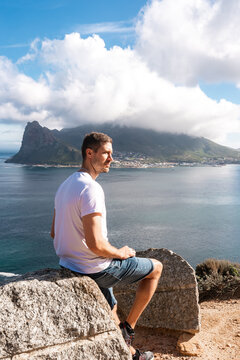 A man in casual clothes sits on a coastal boulder facing Hout Bay and the towering peak under clouds along the Atlantic coast in Cape Town, South Africa.