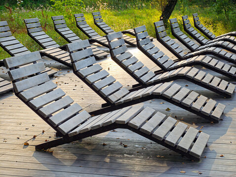 Row of ergonomic outdoor loungers (wave-shaped benches or forest sofas with wooden or polymer-sand slats on a metal frame, set on a wooden decking in the park.