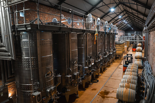 Rows of large stainless steel fermentation tanks and wooden oak barrels line an industrial winery cellar hall with a brick wall and forklift vehicles.
