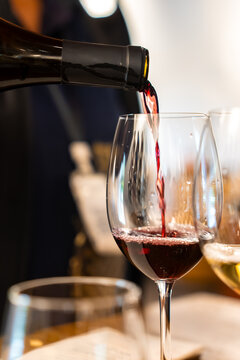 Dark red wine pours smoothly from an open dark bottle neck into a stemmed clear glass during an indoor drink tasting session.