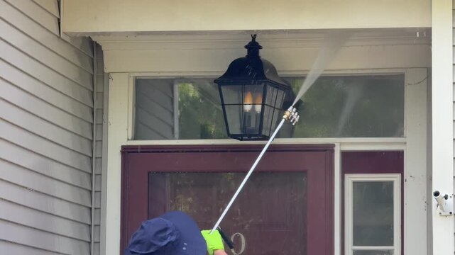 Person with floppy hat and rubber gloves spraying water to clean front entrance to a house of dirt and cobwebs