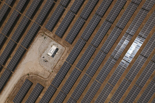 Aerial view of a large solar farm with rows of photovoltaic panels and a small maintenance building on dry ground in Portugal.