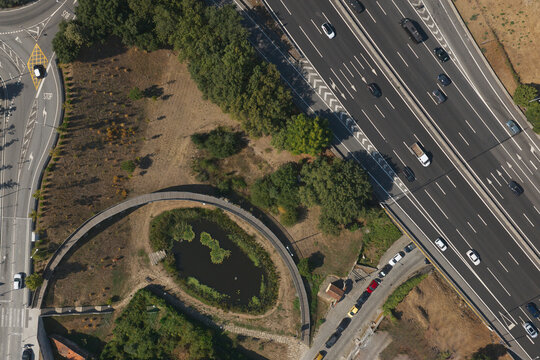 Portugal - 17 August 2026: Aerial view of a small urban lake for water retention, surrounded by a curved walkway, lush trees, and adjacent to a busy multi-lane highway.