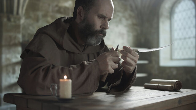 Medieval monk sharpening a quill pen with a knife at a wooden table near a burning candle and scrolls inside a scriptorium