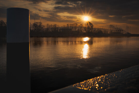 Sunburst and golden reflection on harbor water framed by mooring bollard at sunset in Lauterbach, R&uuml;gen, Germany