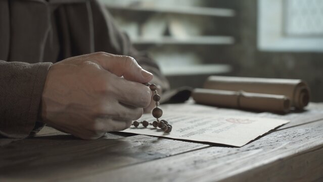 Hands of a monk praying with wooden rosary beads over a manuscript on a rustic wooden table inside a medieval monastery scriptorium