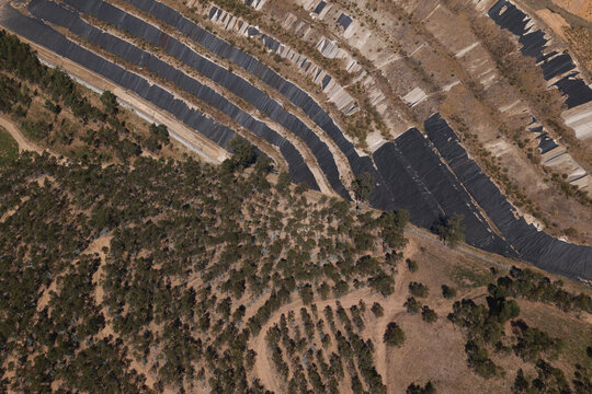Portugal - 14 August 2026: Aerial view of a closed landfill site featuring terraced slopes covered with black plastic sheeting, surrounded by dry vegetation and sparse trees.