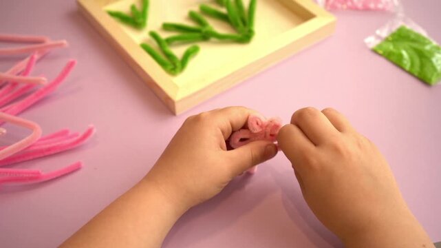 Top view of little kid fingers twisting fluffy pipe cleaners creating handmade artificial blossoms over a wooden frame template for early education and motor skills development