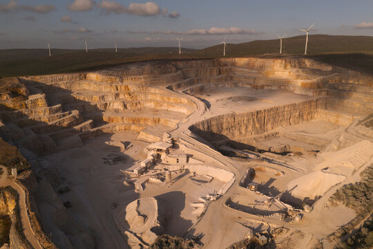 Portugal - 14 August 2026: Aerial view of an open-pit quarry with terraced stone walls, industrial machinery, and wind turbines on surrounding hills under golden hour light.