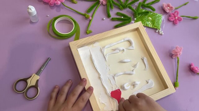 Unrecognizable kid applying sticky paste template with a plastic tool on canvas preparing workspace for handmade pipe cleaner decorations during an early education art class