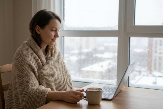 Woman wrapped in a beige blanket sits at a wooden table, using a laptop while holding a cup of coffee, with snow falling outside the window