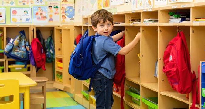 Young boy hanging backpack in kindergarten classroom cubby area
