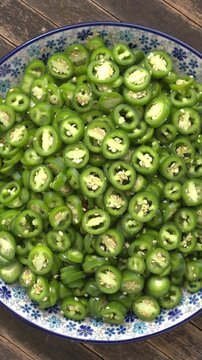 Sliced jalape&ntilde;o peppers pile on a plate. Slowly rotating fresh green chili rings prepared for asian pickles. Backlit by natural window light. Vertical video.