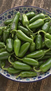 Jalapeno peppers flat lay on plate. Fresh whole green chili heap on floral ceramic dish with dark rustic wood background, slowly rotating in top view with backlit natural window light.