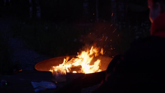 Bright orange flames and glowing embers illuminate rough logs inside round fire pit, set against deep black surroundings with faint smoke, low viewpoint, warm contrast, and intimate gathering mood
