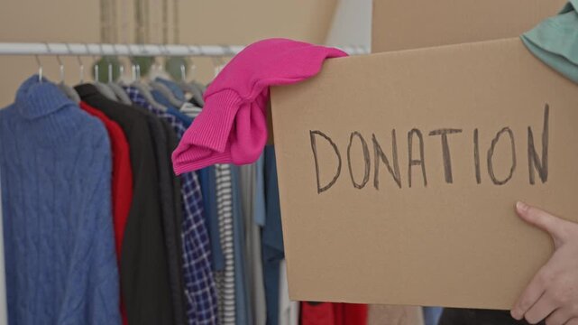 Woman holding a cardboard donation box with hand visible, carrying folded clothes by a clothing rack and shelves in a charity shop; generosity giving.