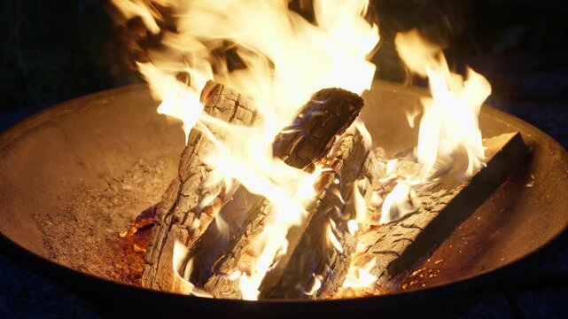 Bright yellow orange firelight fills dark outdoor scene as flames rise around charred logs inside round metal fire pit, with glowing embers, smoky edges, rough bark texture, and cozy leisure mood