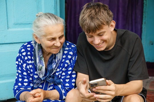 Happy teenage grandson and senior grandmother looking at mobile phone screen, smiling and sharing warm family moment outside.