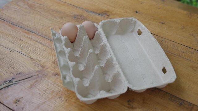 Close-up angled scene shows hand arranging eggs in molded paper carton on scratched wood surface, with warm natural light, blurred green background, practical domestic feel, and sustainable