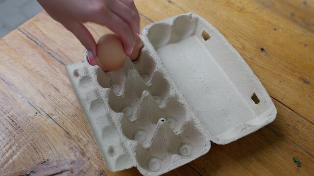 Close-up view shows hand arranging brown and white eggs inside recycled cardboard carton on worn wooden surface, with warm natural light, soft shadows, rough paper texture, and rustic kitchen mood