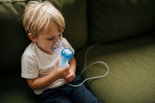 Small blond boy uses nebulizer mask for inhalation therapy at home. Sick child sits on green sofa receiving respiratory treatment for asthma allergy. Pediatric medical care for breathing issues, lung