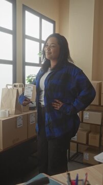 Young woman holding a package with hand on hip and smiling in a small business building packing area; small business optimism.