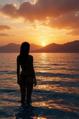 Fototapeta premium Woman in bikini stands in sea water watching sunset over mountains. Orange sky, golden light reflect on ocean waves. Peaceful beach scene, solitary figure enjoys nature.